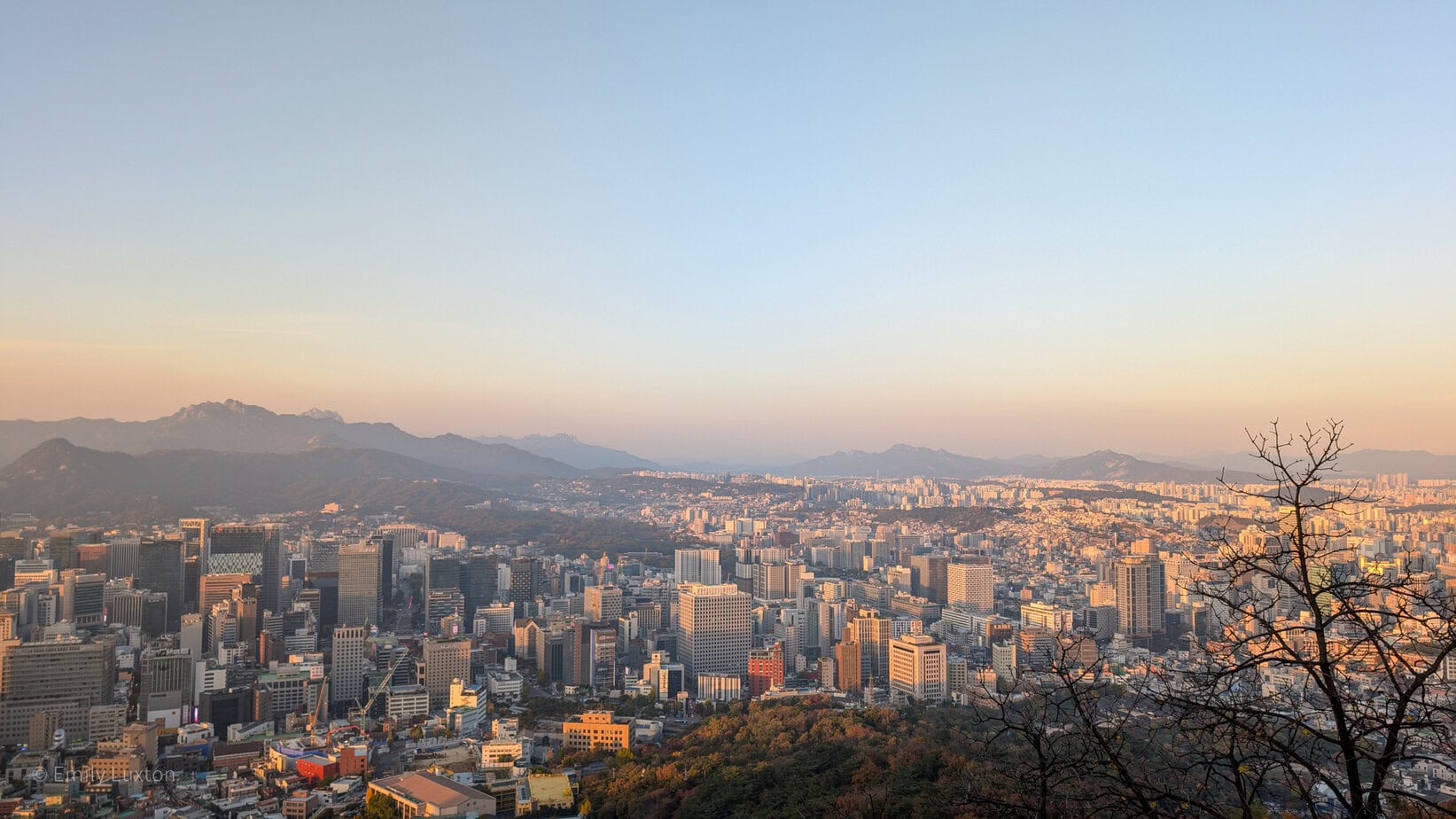 A panoramic view of a cityscape at sunset, featuring a mix of modern skyscrapers and smaller buildings. In the background, mountain ranges are visible under a clear sky with a warm, golden hue. A leafless tree stands on the right side of the image, silhouetted against the setting sun.