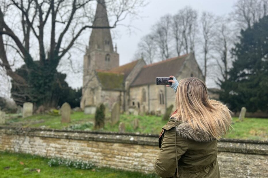 Emily wearing a khaki green parka with a fur lined hood facing away from the camera using a smartphone to take a photo of a church in England on a grey day