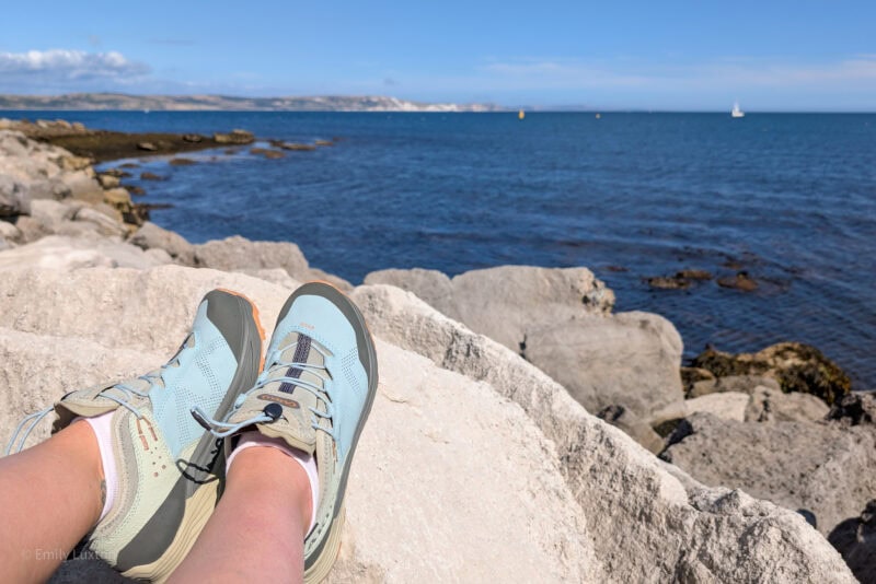 Feet in light cyan coloured hiking shoes propped on a rock with the sea behind on a coastal hiking trail in summer.