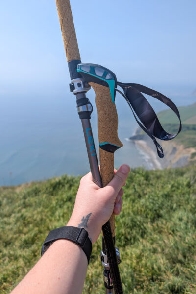 Hand holding two trekking poles in front of a view of cliffs and blue sea on a sunny summer day
