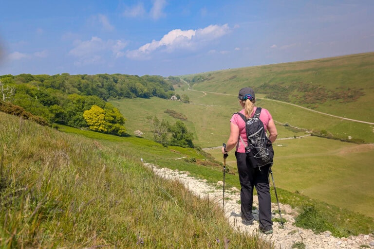 Emily hiking on a gravel path overlooking a grassy valley. She is wearing black trousers, a pink top, and a black backpack and using trekking poles. UK summer hiking essentials.
