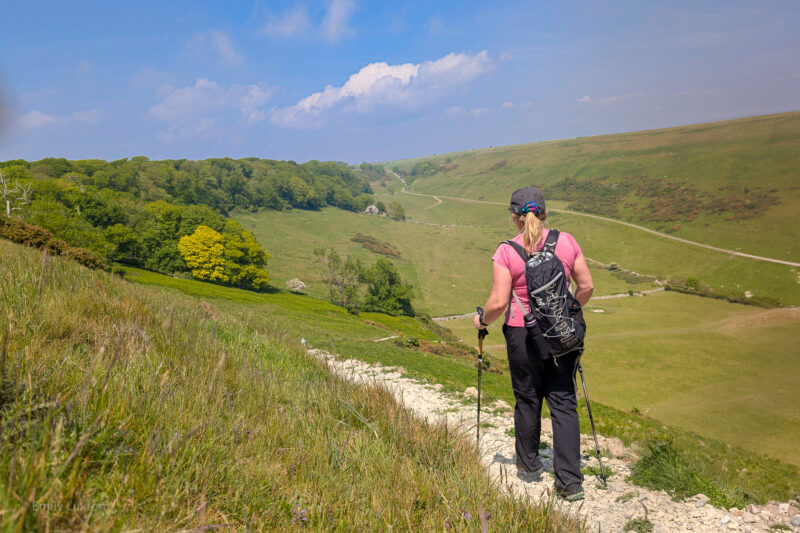 Emily hiking on a gravel path overlooking a grassy valley. She is wearing black trousers, a pink top, and a black backpack and using trekking poles. UK summer hiking essentials.