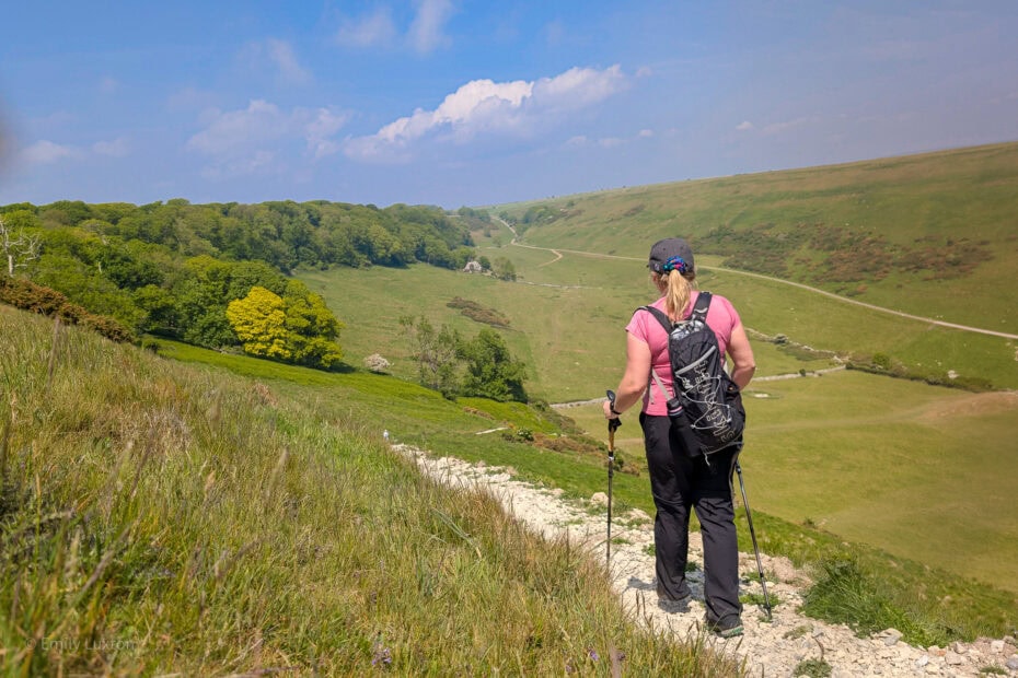Emily hiking on a gravel path overlooking a grassy valley. She is wearing black trousers, a pink top, and a black backpack and using trekking poles. UK summer hiking essentials.