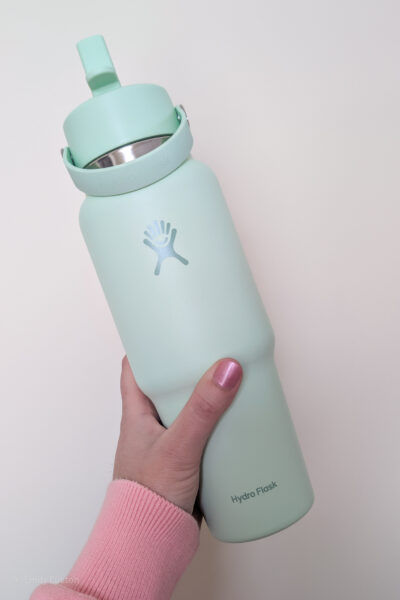 Hand holding a large mint green Hydro Flask bottle in front of a cream wall.
