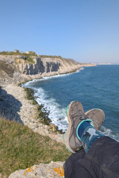 Feet in blue socks and grey shoes on a cliff edge with grey rocky cliffs and blue sea in the background