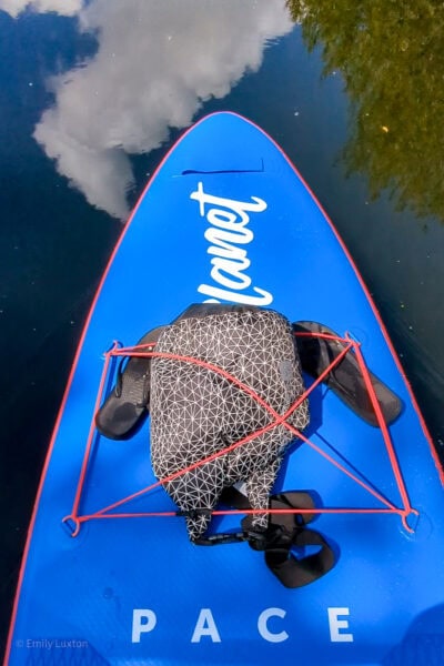 blue paddleboard on a river with a black and white dry bag and black flip flops on top
