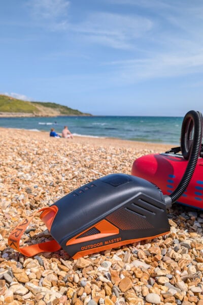 Black and orange electric pump with the name Outdoor Master on the side, next to a red paddleboard on a stony beach with the sea in the background