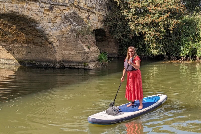 Emily in a red dress and grey life jacket standing on a blue and white paddleboard in a river in front of a stone bridge