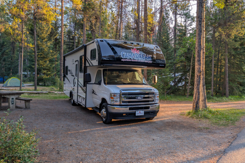 A large motorhome with the CanaDream logo above the cab, parked in a campground plot surrounded by trees