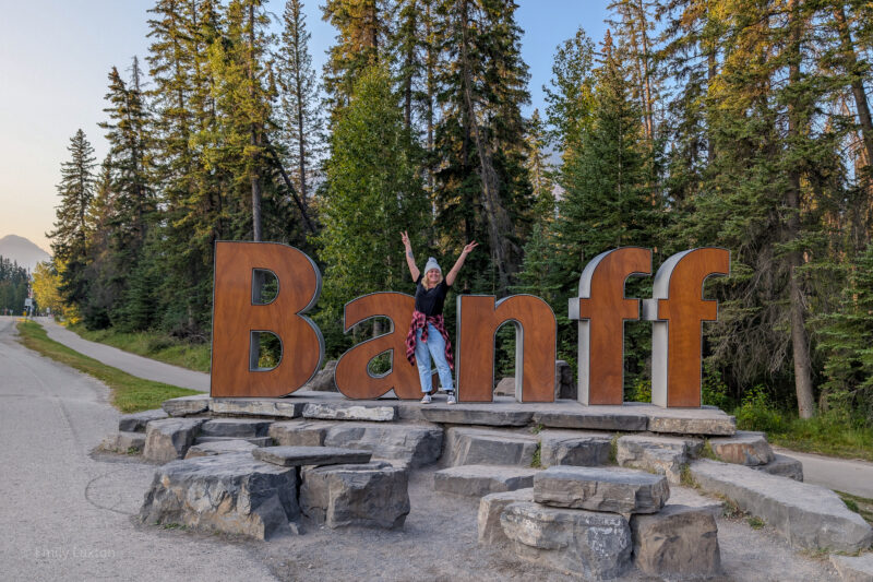 Emily standing in front of a large wooden Banff sign with her hands in the air