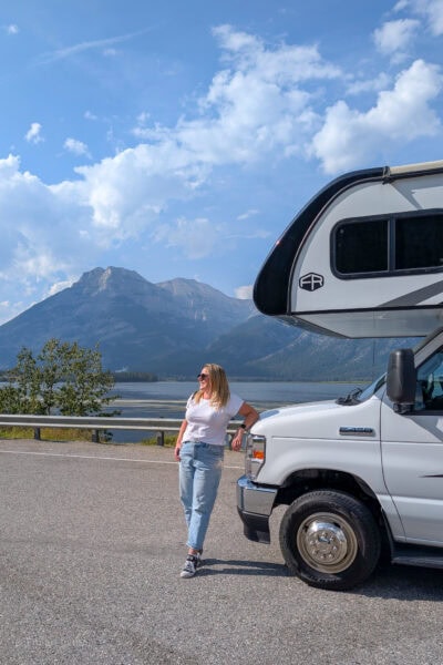 Emily wearing jeans and a white t-shirt leaning on the front of a white motorhome with a lake and mountains behind | Alberta 1 week Road Trip