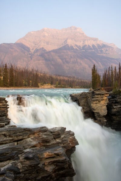 Large, forceful waterfall with a mountain behind lit in pink at sunset
