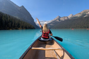 Emily in the prow of a wooden canoe on a very blue lake with her hand in the air and mountains behind