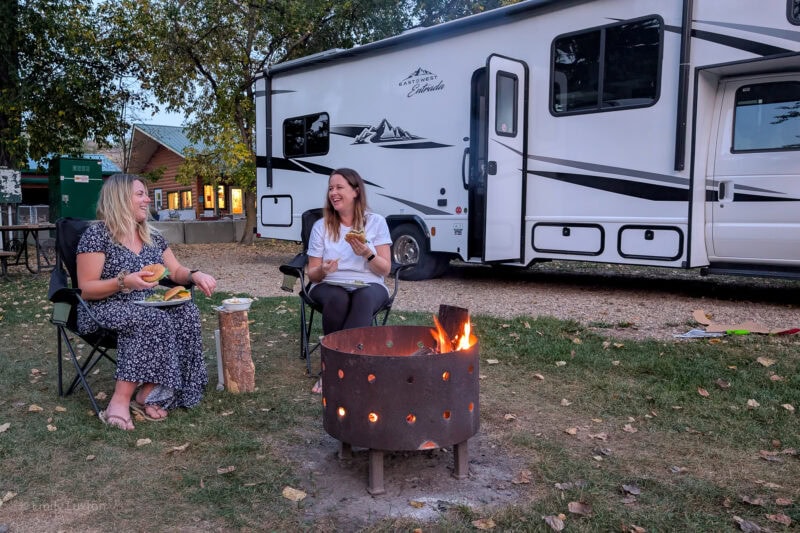 Two girls sitting on camping chairs by a small campfire with a large white motorhome behind