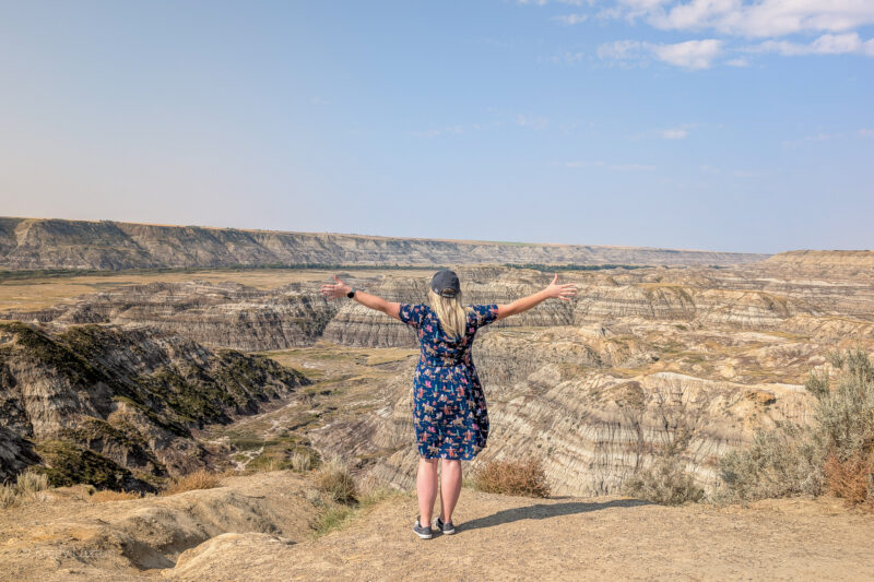 Emily in front of a dry landscape with layered sandstone pillars. She is wearing a blue dress nad has her arms out wide.