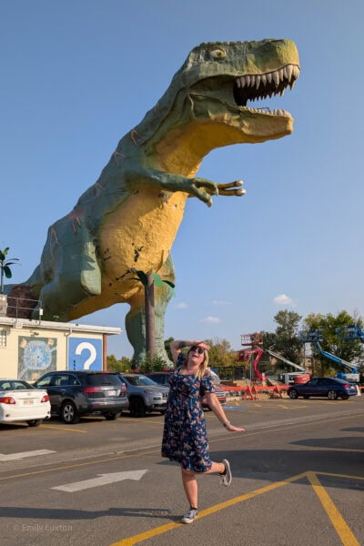 Emily posing in front of a very large green t-rex statue with one hand on her head like a damsel in distress