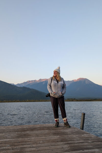 Emily wearing a grey hoodie standing in front of a lake with mountains behind