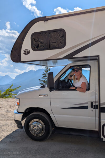 White motorhome with mountains behind and emily leaning out the window wearing a grey cap