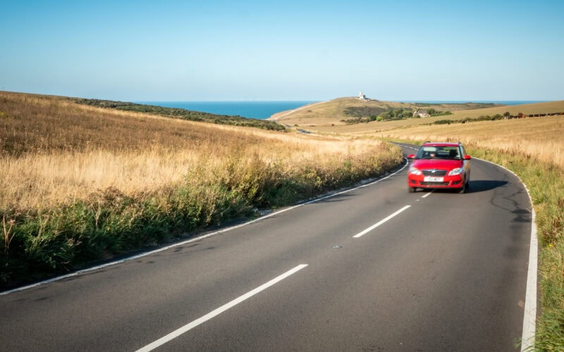 A car travelling a winding road on the South Downs (East Sussex, England) with Belle Tout lighthouse and the English Channel in the distance.