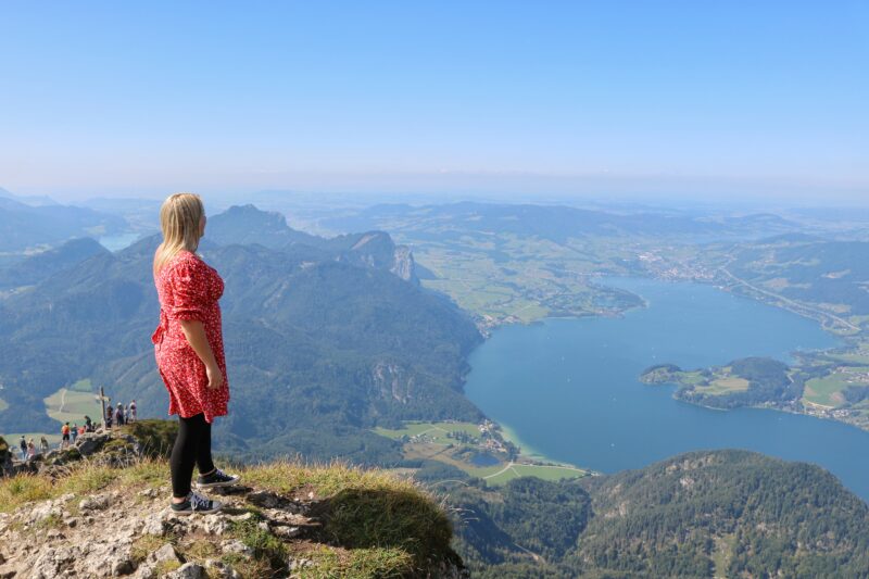 Travel blogger Emily Luxton in a red dress and black leggings stands on a rocky outcrop, looking out over a large body of water surrounded by green hills and mountains with clear blue sky overhead.
