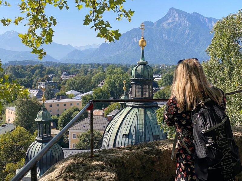Emily taking a photo of Salzburg skyline with a green dome in front of her and mountains in the background