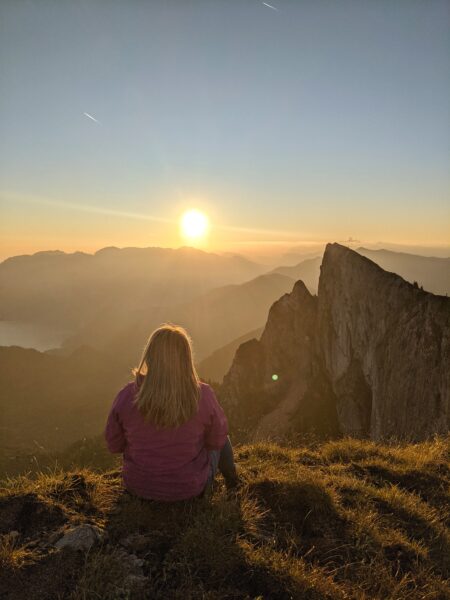 emily in a pink jacket on a mountain top facing a jagged peak at sunrise
