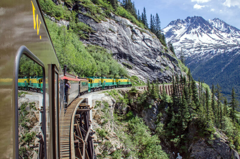 Looking out a train window as the train curves along a cliff next to a mountain