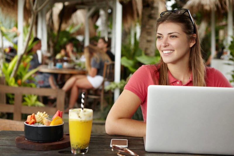 Happy young self-employed woman enjoying free wireless internet connection sitting in front of generic laptop at outdoor cafe. Joyful female using notebook computer during lunch at sidewalk restaurant