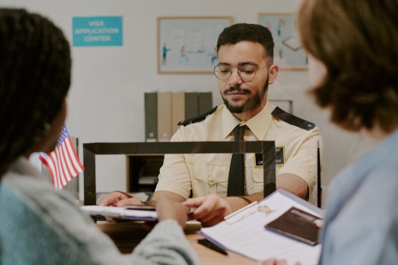 Officer processing visa application for two individuals at counter with American flag present and documents in hand. Focus on customer interaction and professional setting