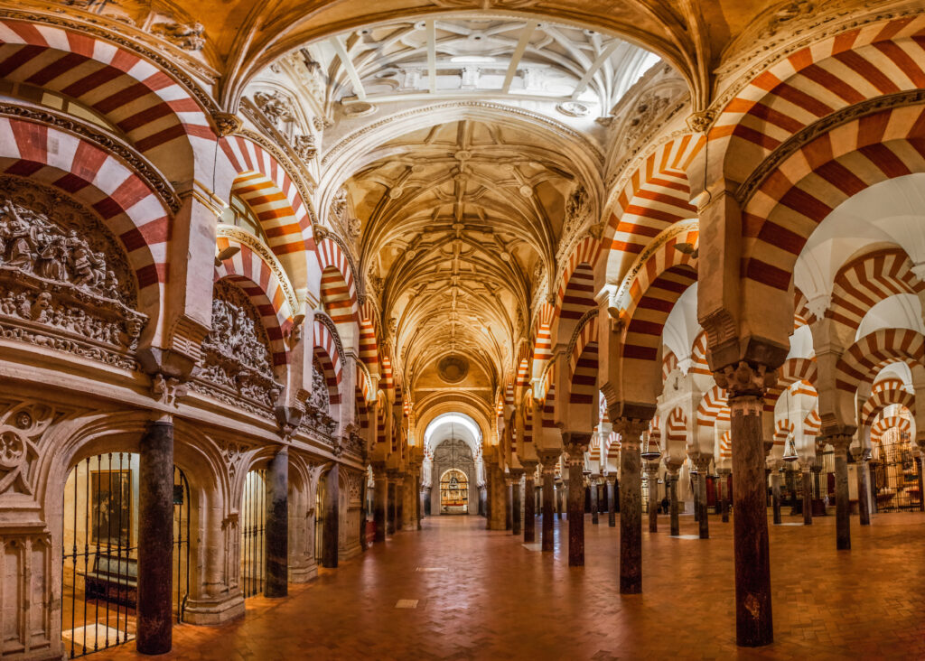 Interior of the Mosque-Cathedral of Córdoba showing red-and-white arches, stone columns, and intricate vaulted ceilings in a blend of Islamic and Christian architecture.