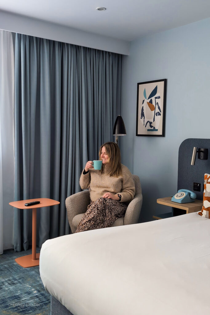 emily sitting in an armchair drinking coffee from a blue mug. she is wearing a beige jumper.