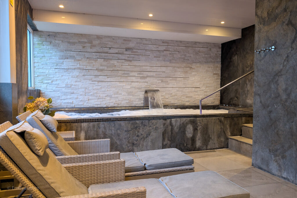 interior of a spa with grey tiled walls. There are lounge beds and a large hydro pool behind.