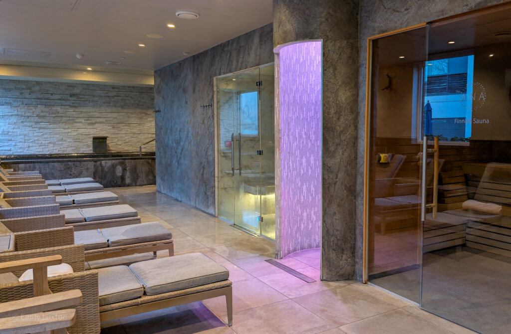 interior of a spa with grey tiled walls. There are lounge beds facing a sauna with glass walls.