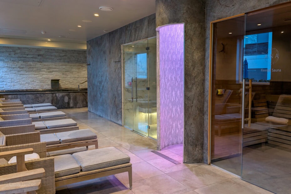 interior of a spa with grey tiled walls. There are lounge beds facing a sauna with glass walls.