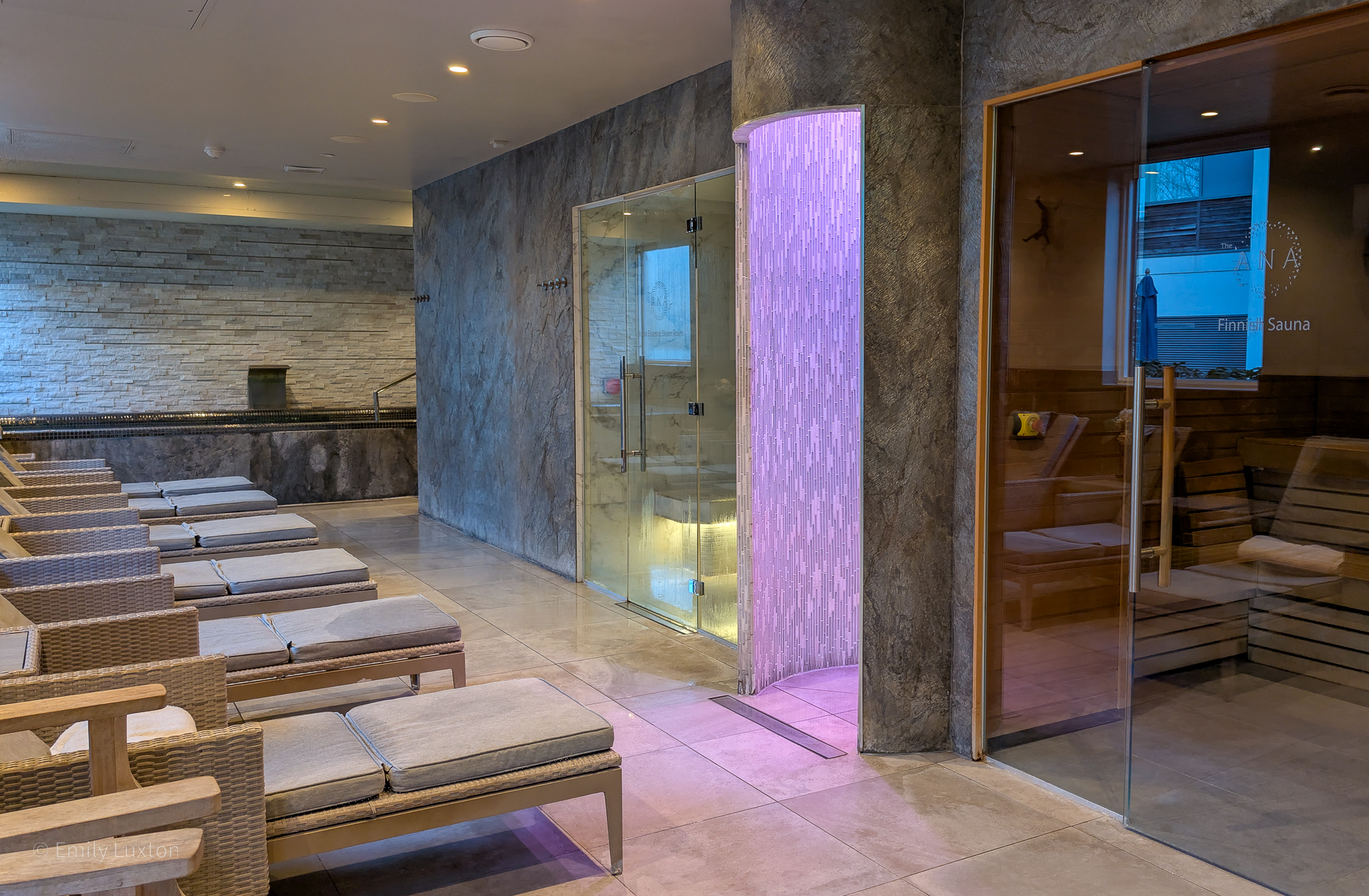 interior of a spa with grey tiled walls. There are lounge beds facing a sauna with glass walls.
