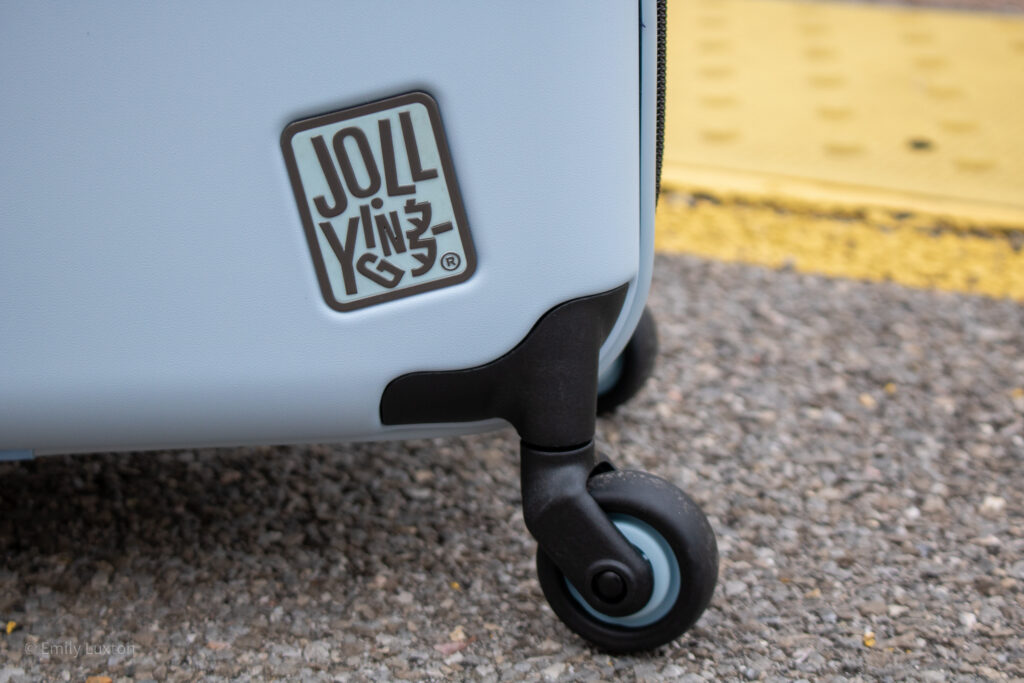 Close up of the bottom right corner of a light blue hard shell suitcase with the logo JOLLYING and black wheels