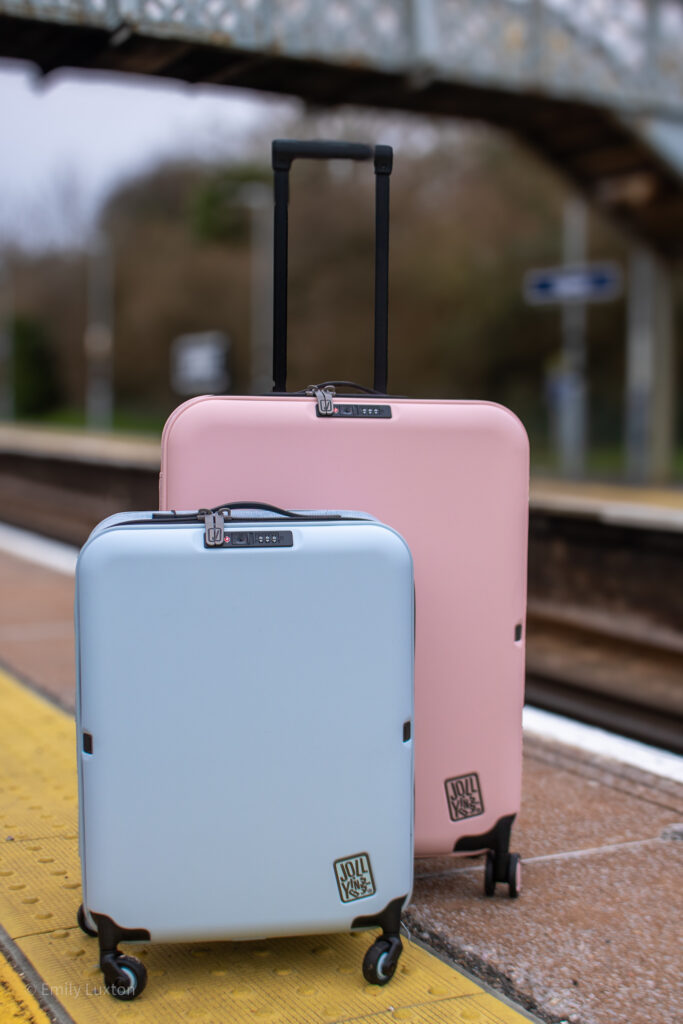 A large pink and small blue hardshell suitcase on a train station platform with an iron bridge behind | JOLLYING Pebble Review