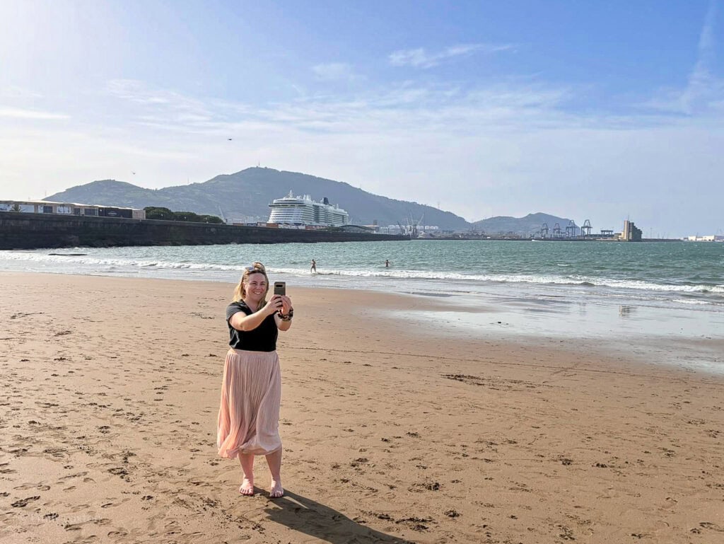 emily on a beach wearing pink skirt and black t-shirt taking a photo with her phone
