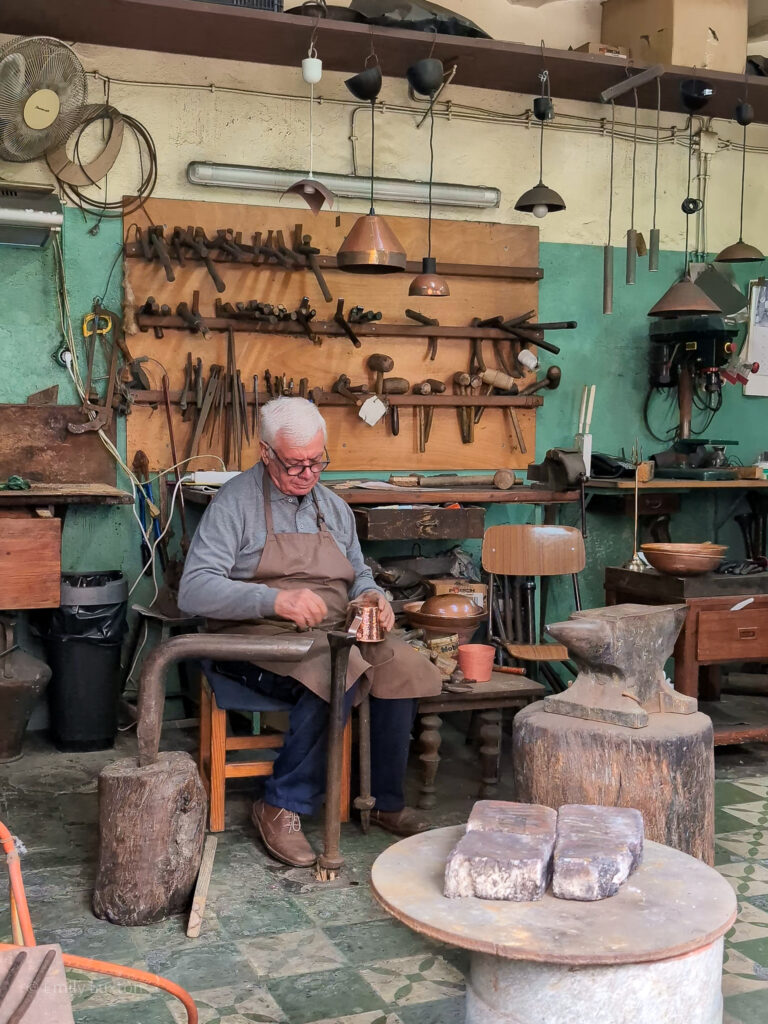 Man in a brown leather apron sitting on a stool hammering a copper mug