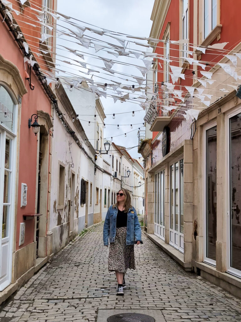 Emily wearing a black t shirt, denim ajcket and leopard print skirt walking on a cobbled street below fluttering white bunting