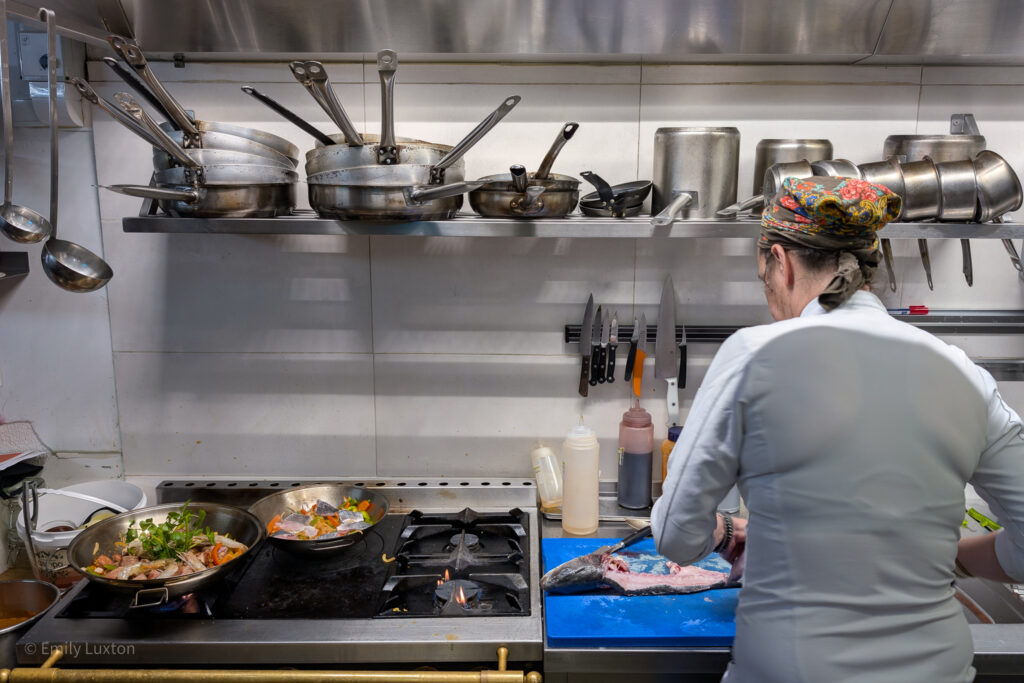 Professional kitchen with a chef gutting a fish next to a hob with two pans full of sliced peppers