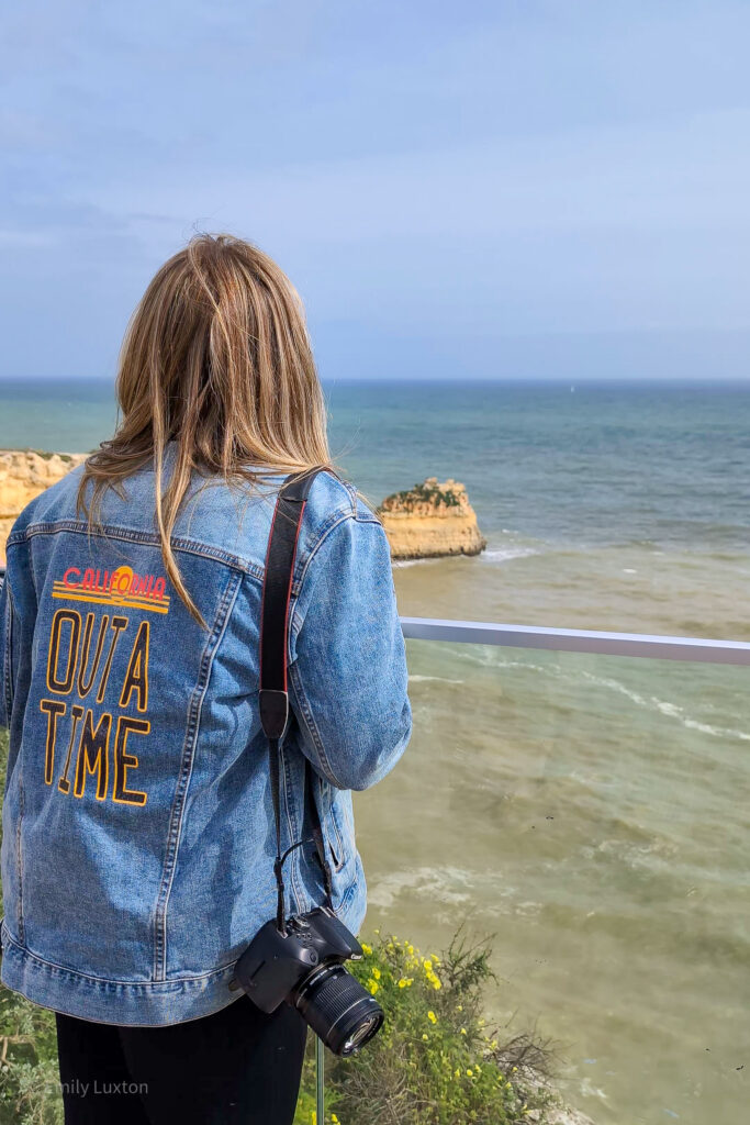 emily leaning against a glass railing looking out to sea towards an orange coloured rock stack. She is wearing a blue denim jacket with OUTA TIME printed on the back.