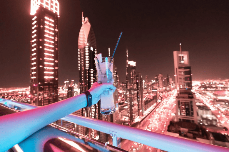 hand holding a cocktail over a blacony at night with dubai city skyline behind