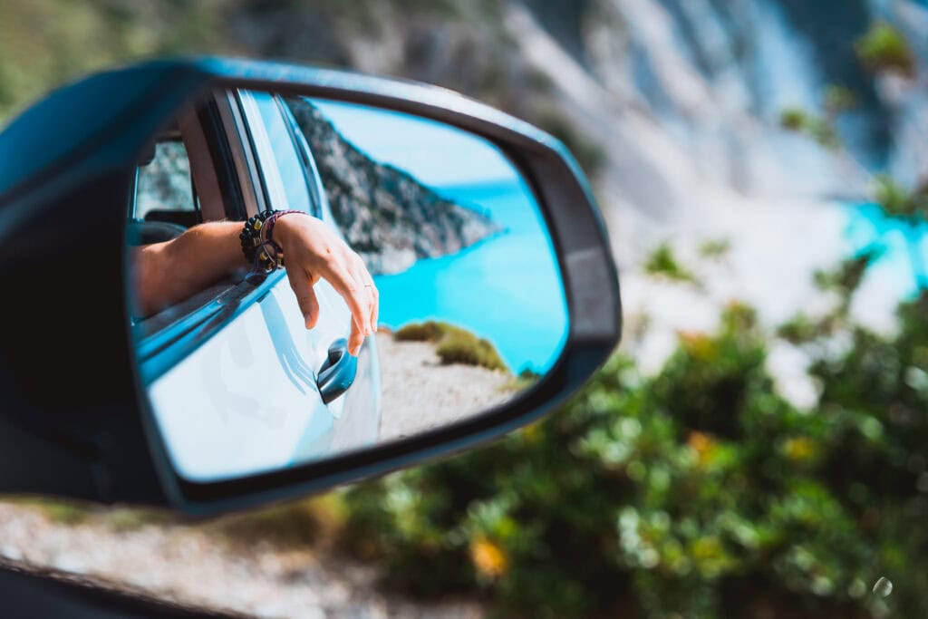 Female hand mirrored in the car side view mirror. Blue mediterranean sea and white rocks in background.