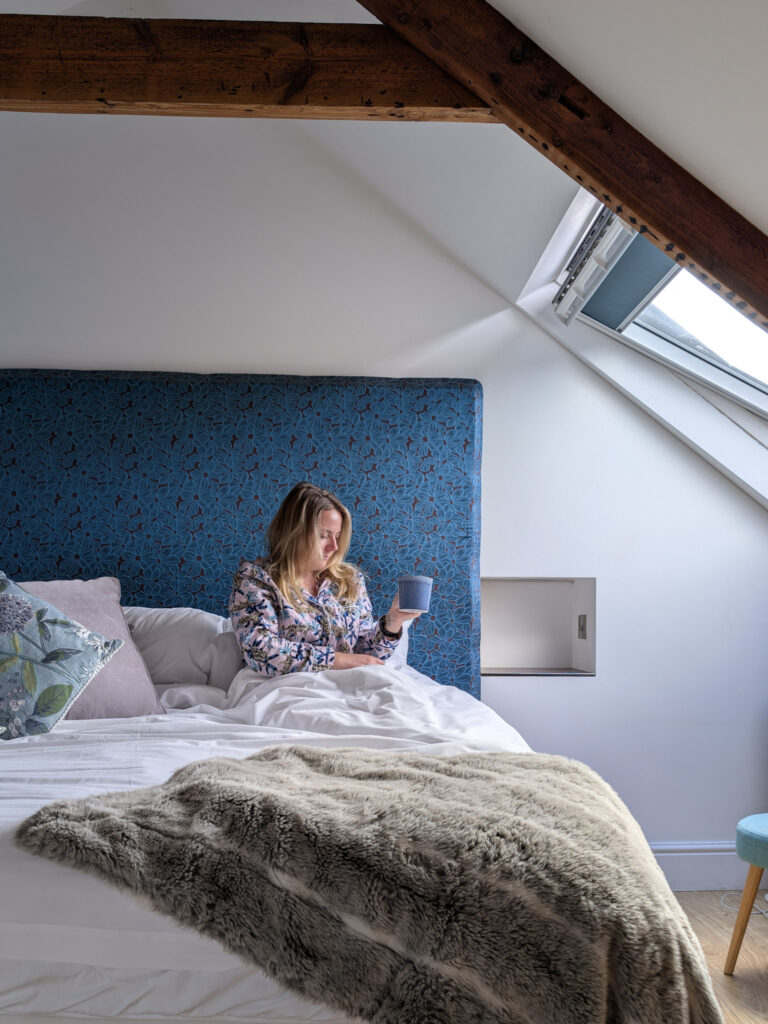 Emily in bed wearing pink pyjamas and holding a blue coffee mug, there is a blue headboard behind