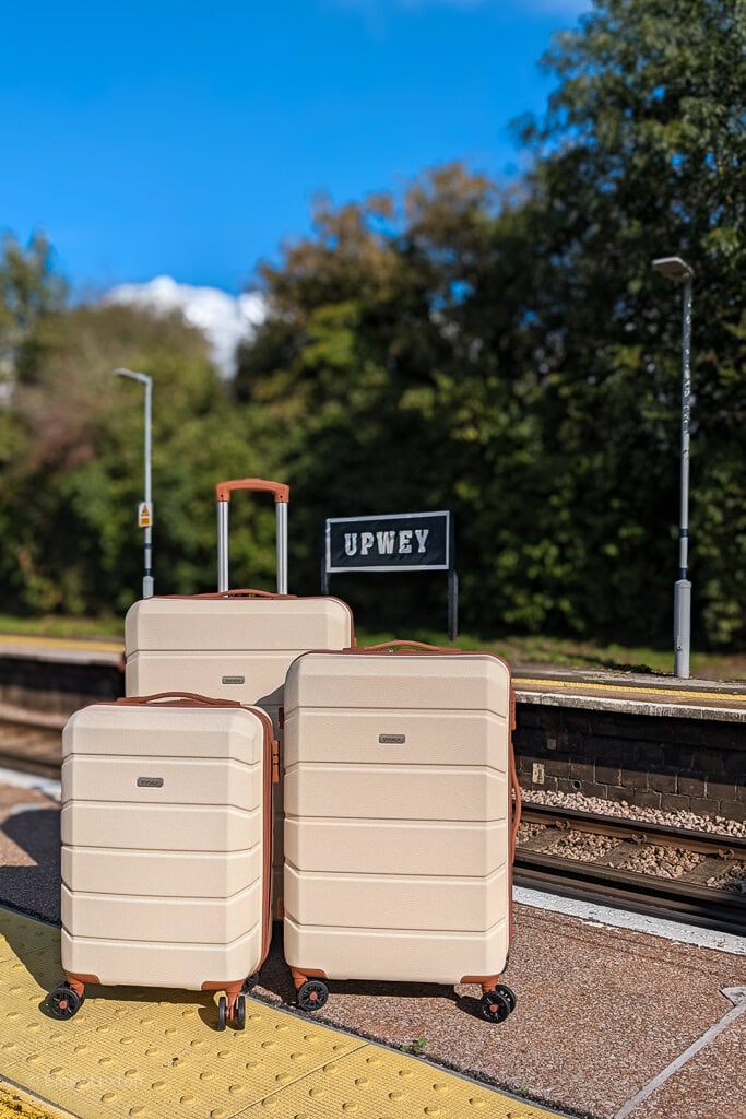 3 cream suitcases with brown trim on a train station platform with a sign in the background reading 'Upwey'