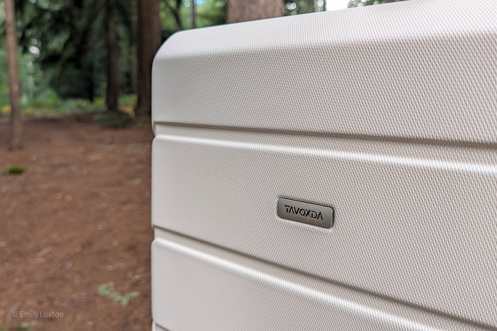 Close up of the front corner of a cream hard shell suitcase with a silver label marked 'TAVOXDA'