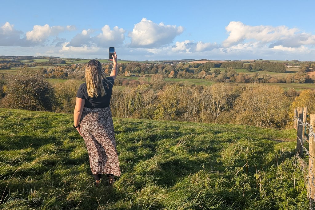 Woman wearing a black t-shirt and leopard print skirt standing in a field taking a photo on a phone